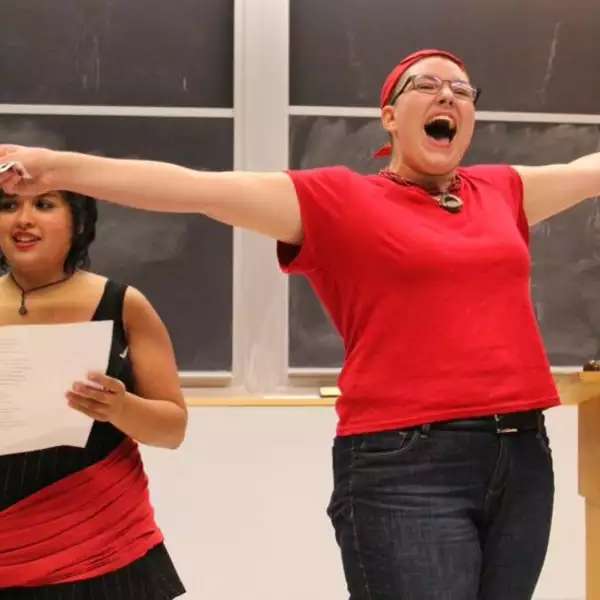 Two students presenting their work in a classroom. One student to the left holds a sheet of paper and looks towards the class. The other student to the right has her arms spread out and her mouth wide open singing or shouting.