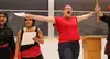 Two students presenting their work in a classroom. One student to the left holds a sheet of paper and looks towards the class. The other student to the right has her arms spread out and her mouth wide open singing or shouting.